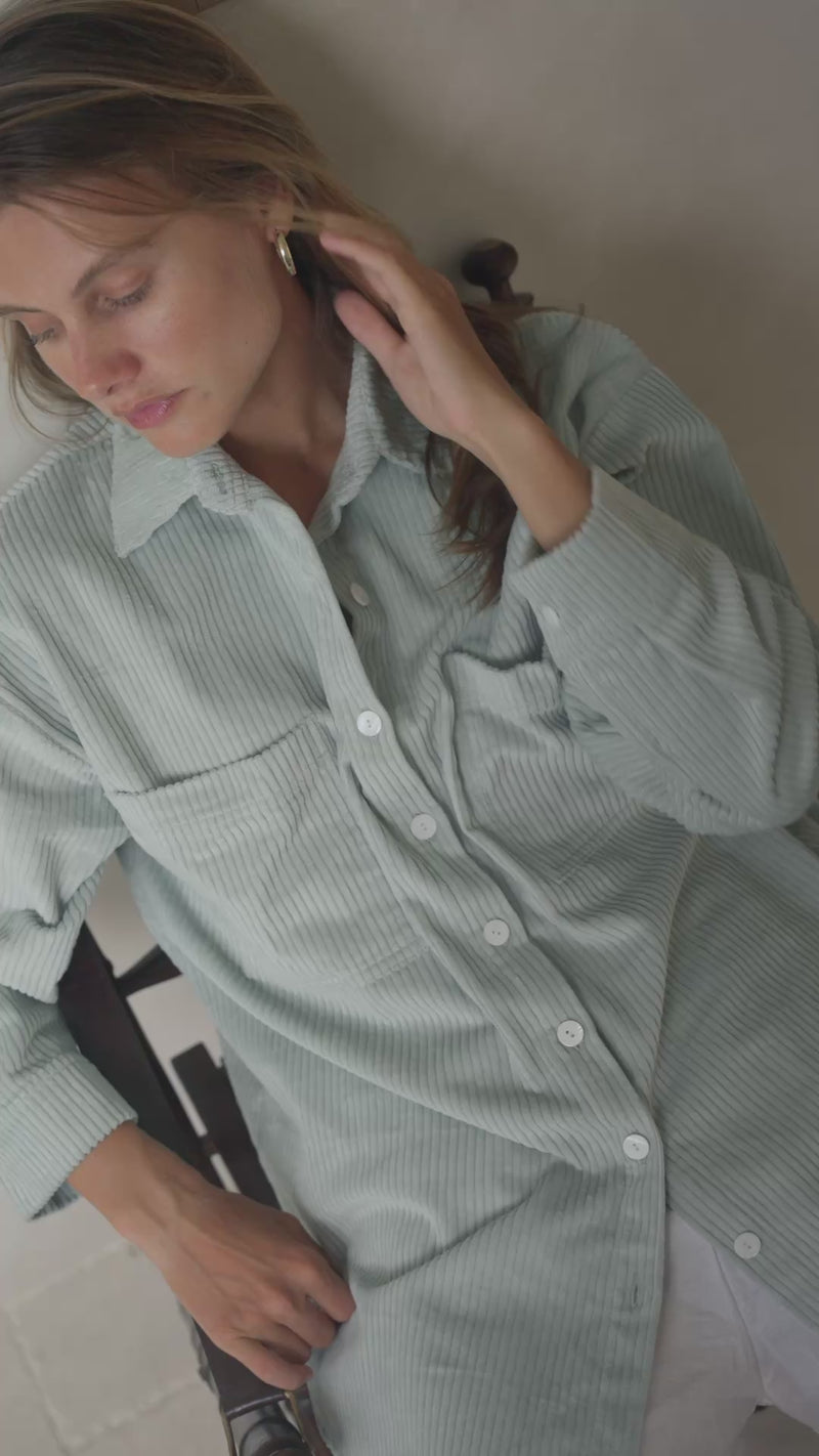 Woman sitting in a softly lit room wearing a light green corduroy jacket and white shorts.