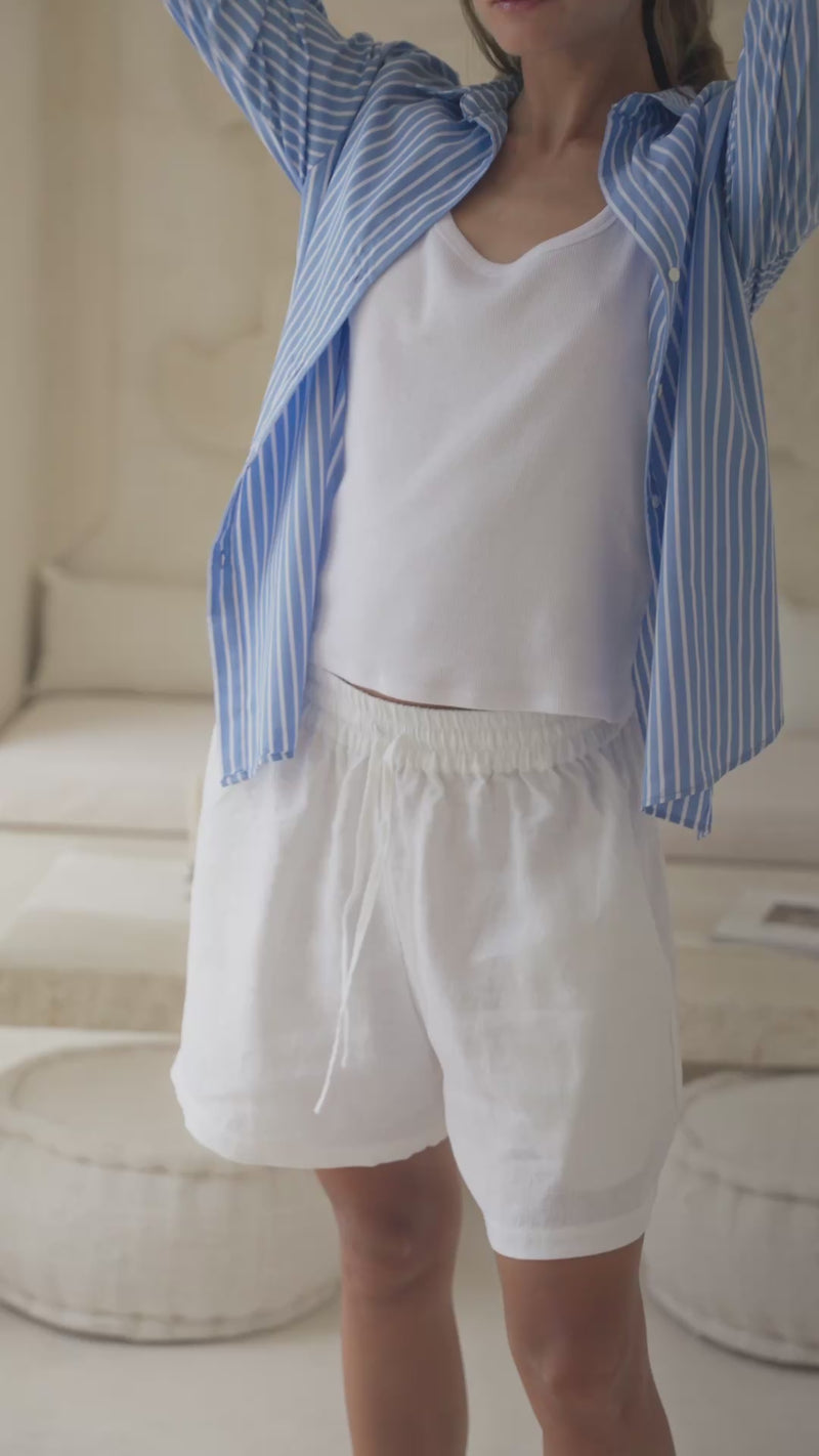 Woman in a blue striped shirt over a white tank top and white shorts standing in a stylish living room.