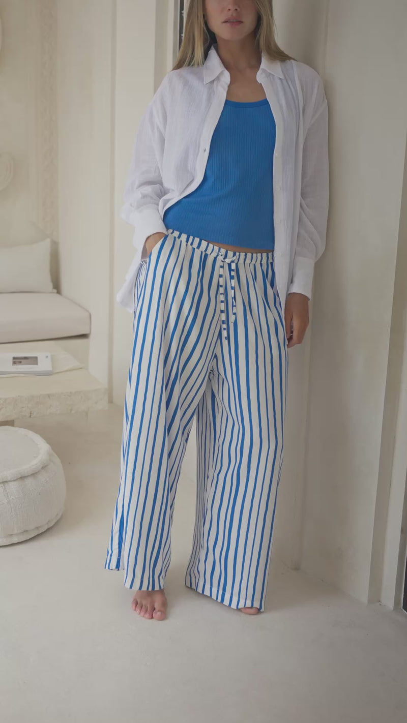 Woman wearing a white shirt, blue top, and striped pants standing in a softly lit room.