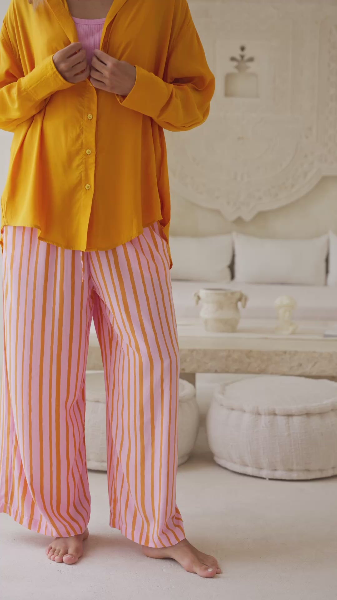 Load video: Woman wearing an orange shirt over a pink top and orange striped pants standing next to a large white wall.