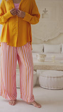 Load and play video in Gallery viewer, Woman wearing an orange shirt over a pink top and orange striped pants standing next to a large white wall.
