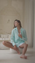 Load and play video in Gallery viewer, Woman in a light blue shirt and white shorts sitting on a white ottoman in a bright room.

