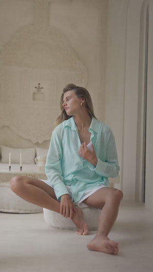 Woman in a light blue shirt and white shorts sitting on a white ottoman in a bright room.