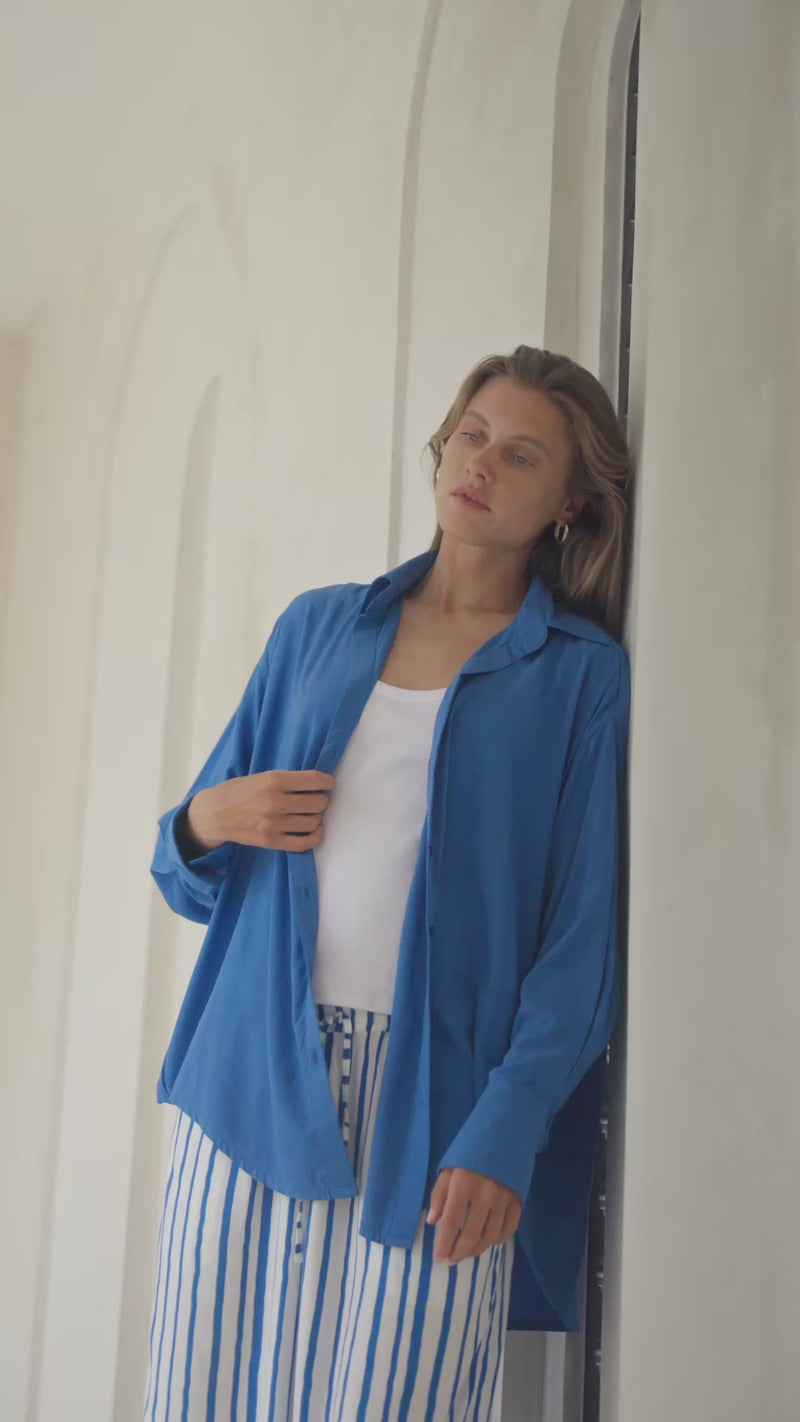 Woman standing by an arched doorway wearing a blue shirt, white tank top, and striped paants.
