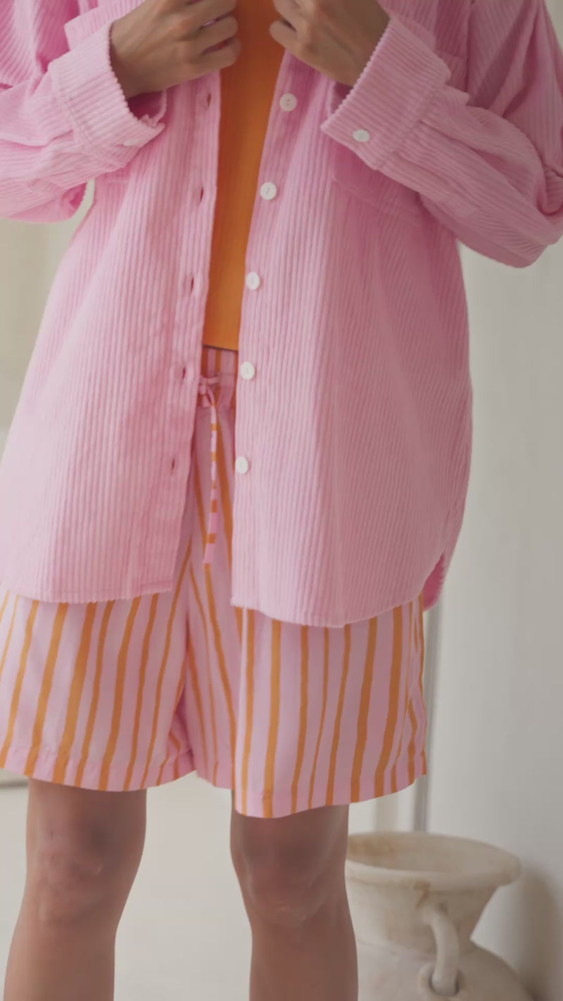 Woman wearing a pink corduroy shirt over an orange top and striped shorts, standing in a light and airy room