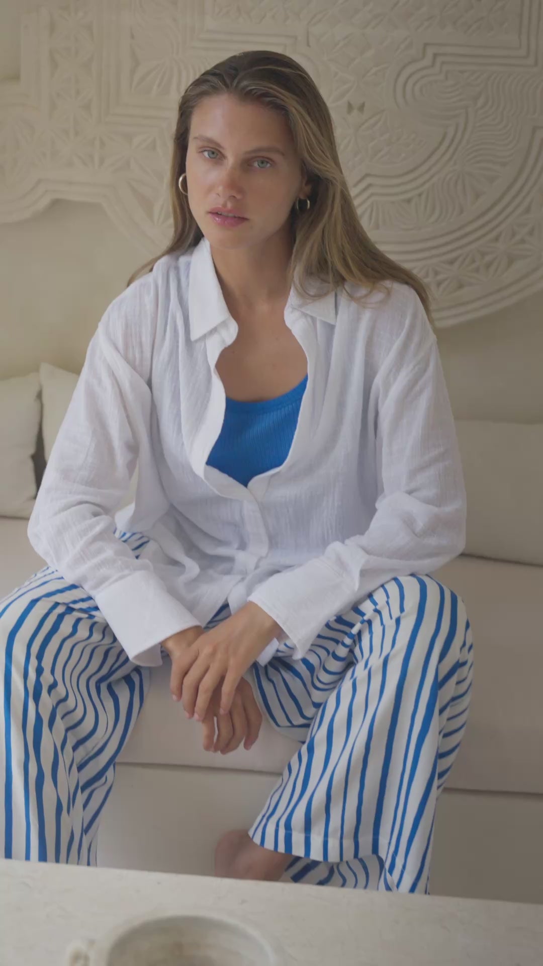 Load video: Woman wearing a white shirt, blue top, and striped pants sitting in a softly lit room.