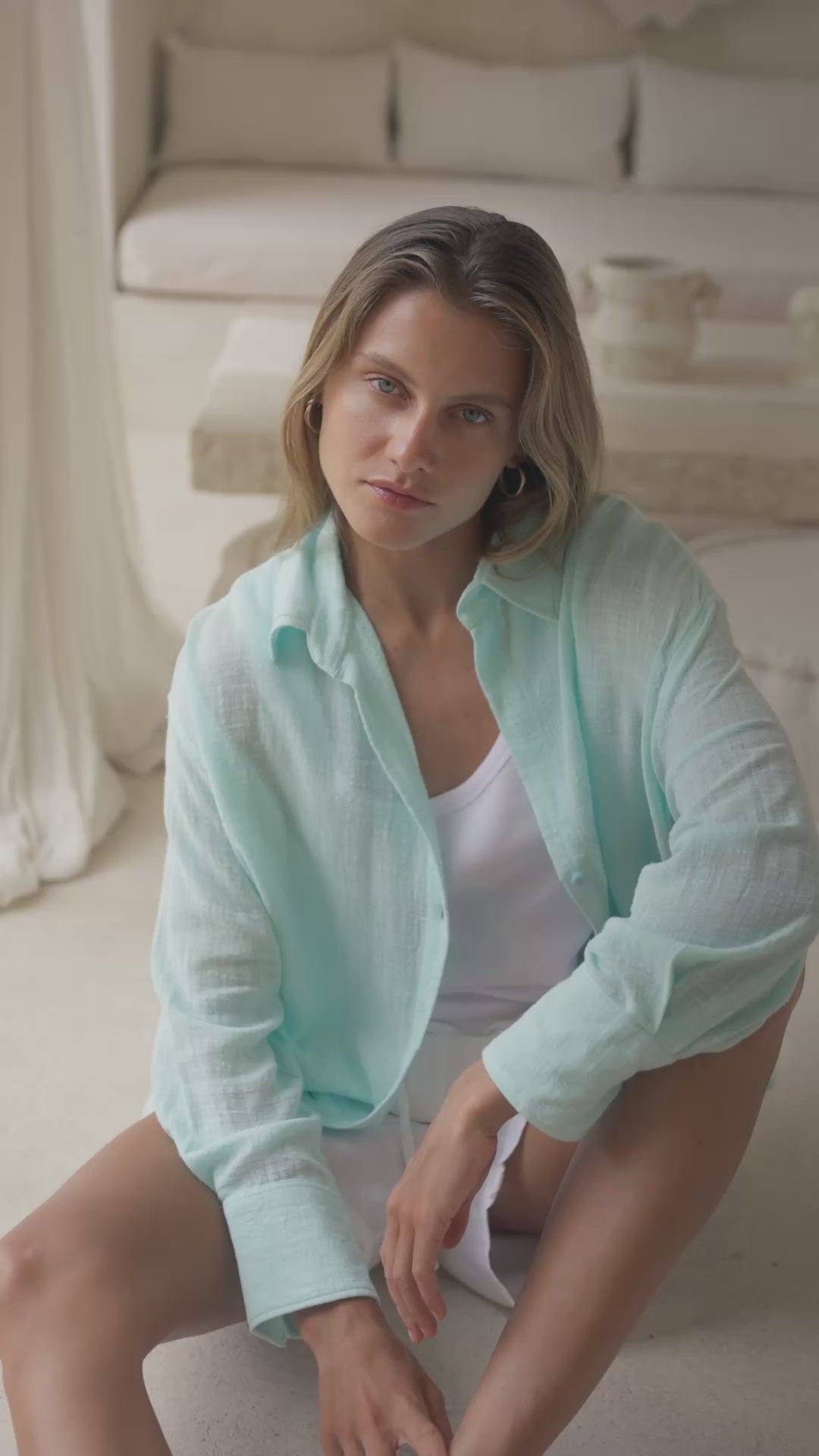 Load video: Woman in a light blue shirt and white shorts sitting on a white ottoman in a bright room.
