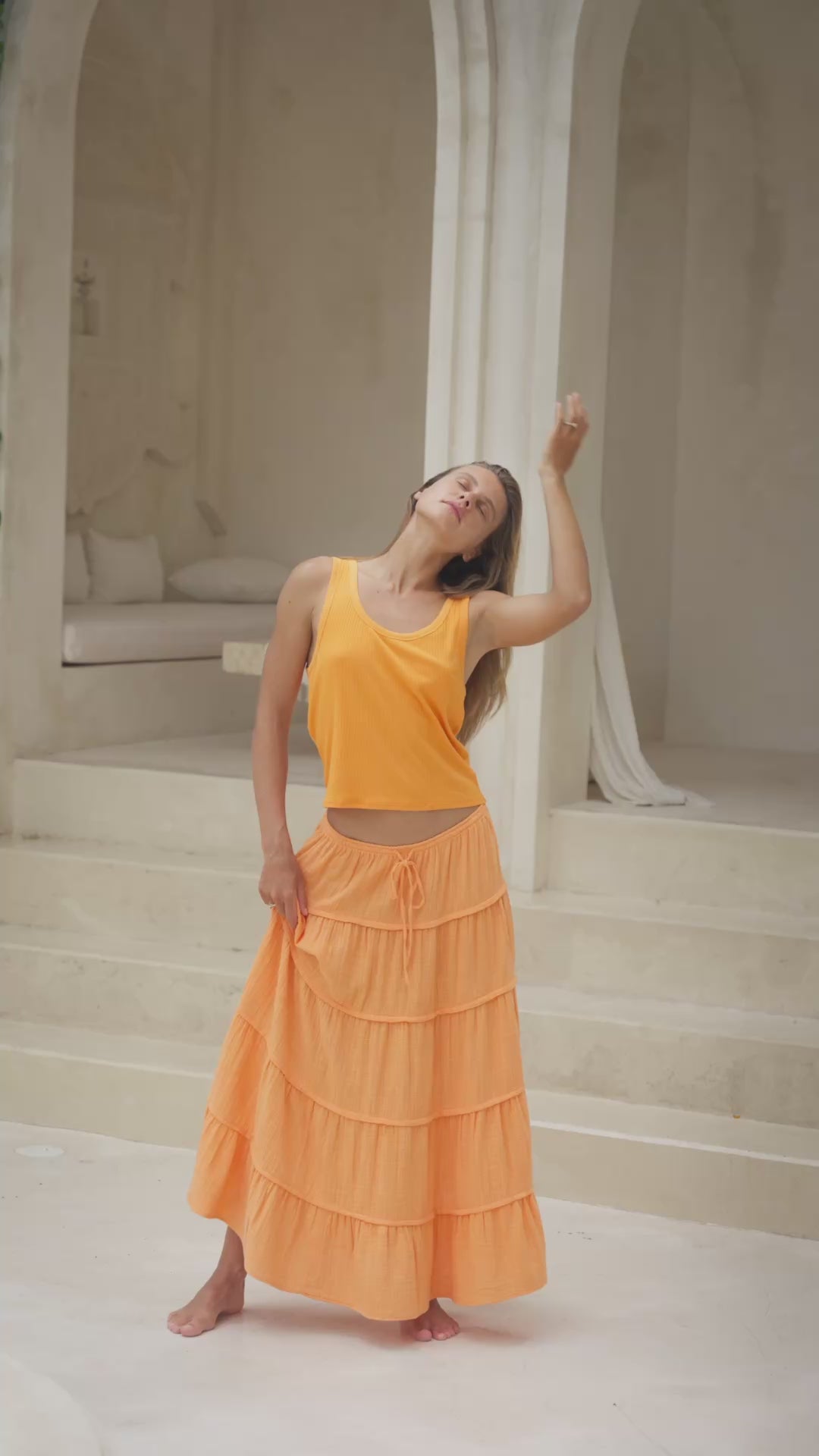 Load video: Woman in an orange skirt and tank top standing in front of a classical architectural archway and steps.