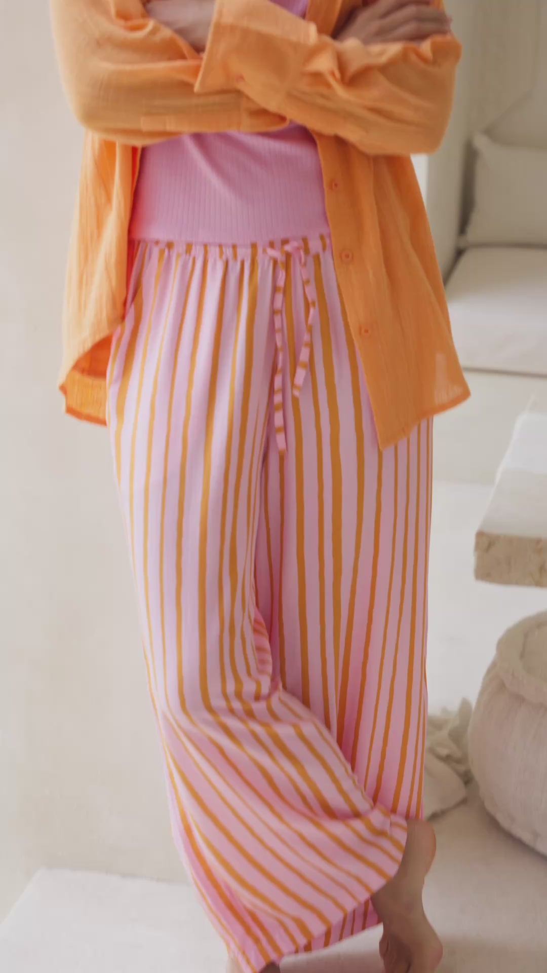 Load video: Woman wearing an orange shirt and pink striped pants in a bright room.