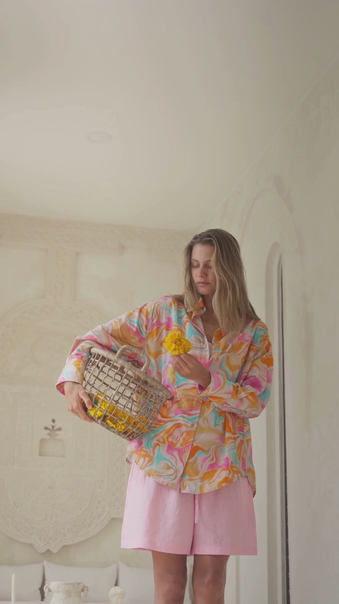 Load video: Woman wearing a colorful patterned shirt in a bright room with a basket of flowers