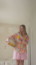 Load and play video in Gallery viewer, Woman wearing a colorful patterned shirt in a bright room with a basket of flowers
