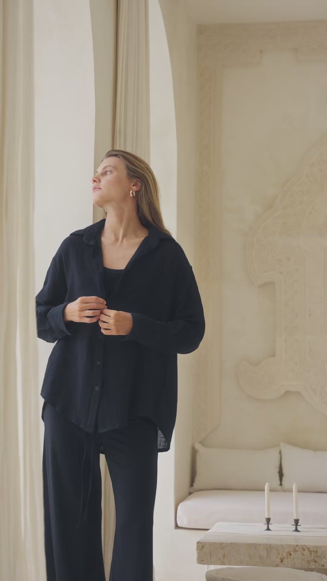Load video: Woman in a black shirt, tank top and pants standing in a luxurious room with white decor