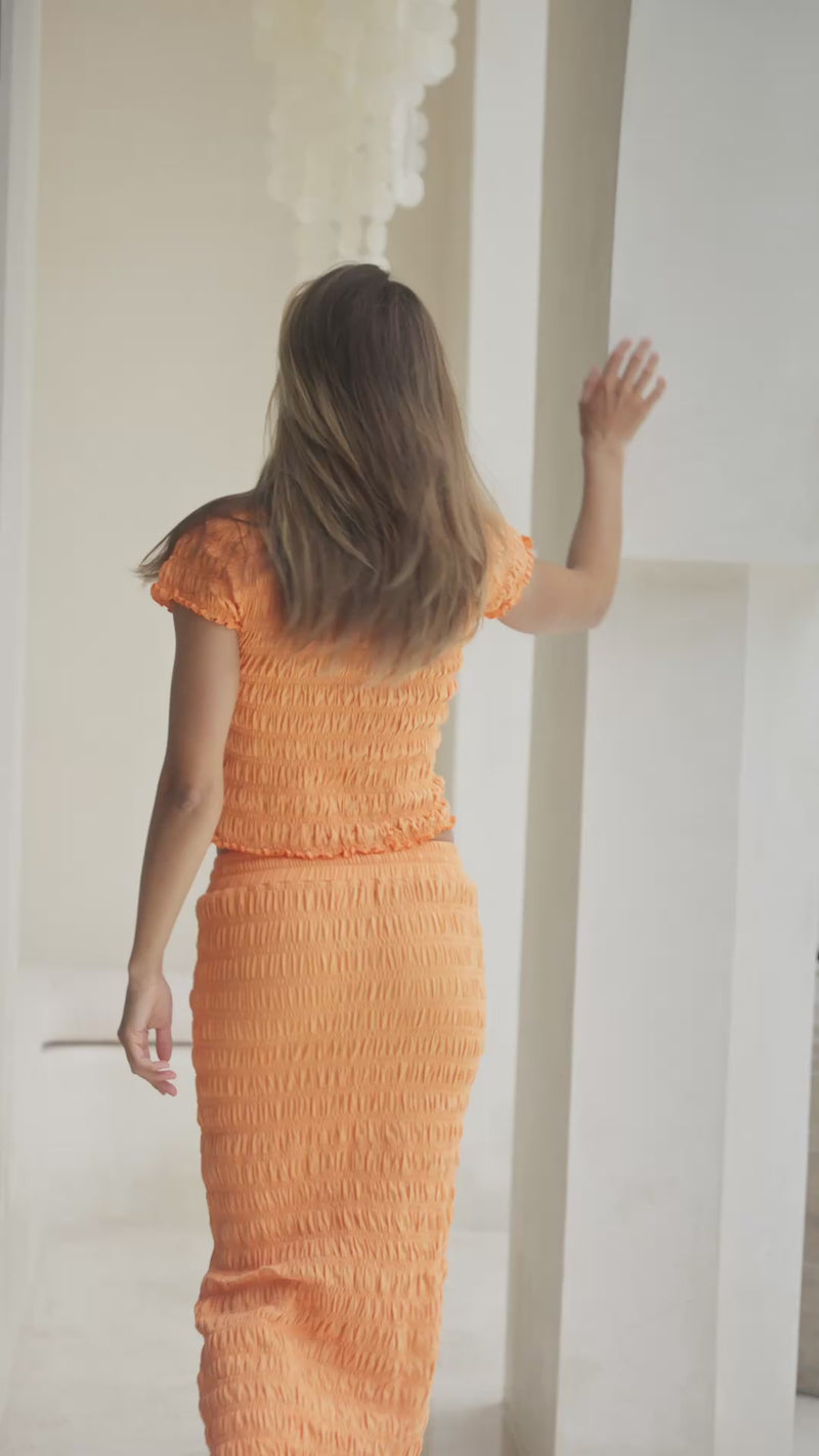 Woman in orange shirred top and maxi skirt standing on steps with a decorative wall in the background