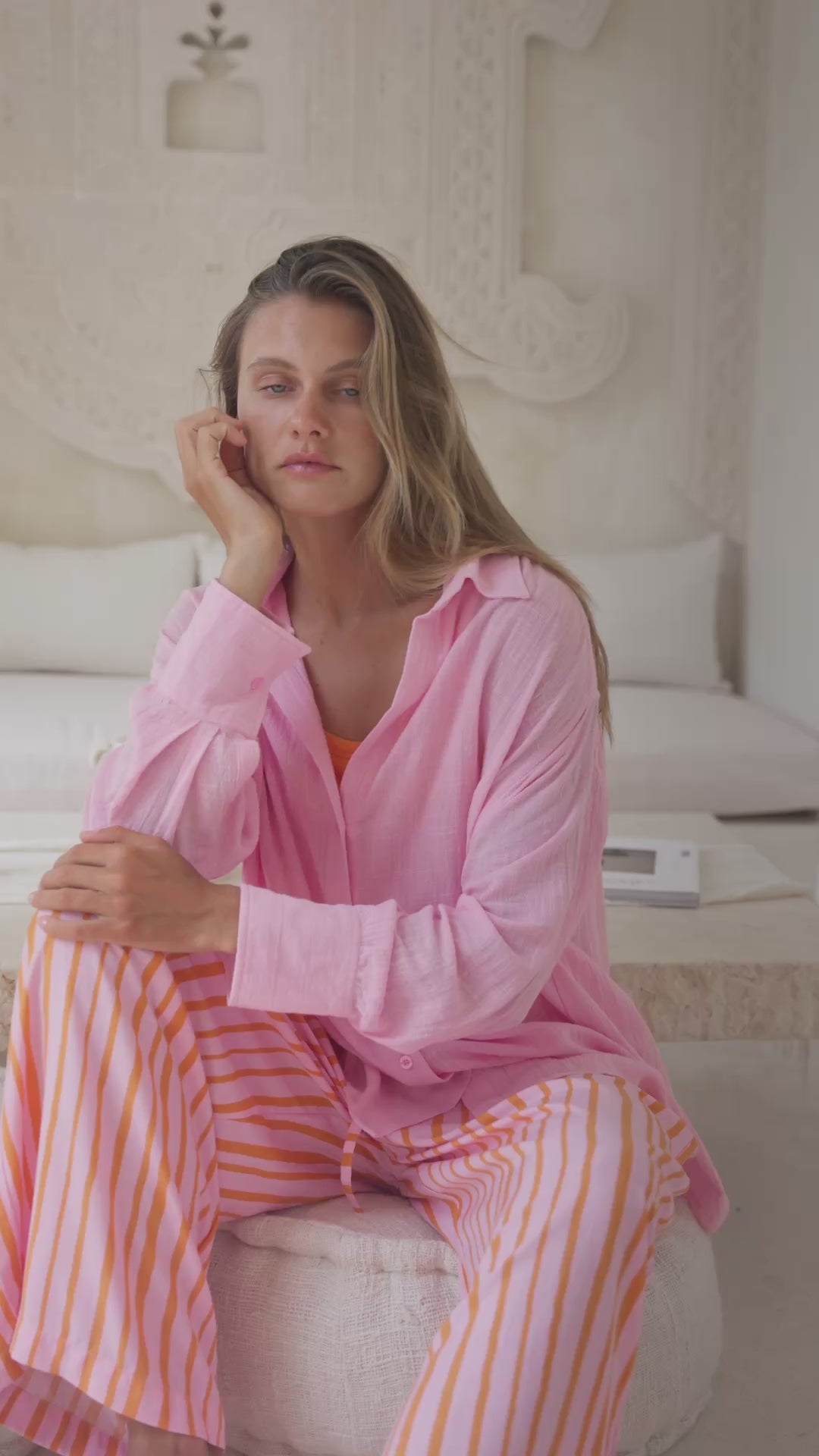 Load video: Woman wearing a pink shirt over an orange top and striped pants sitting on an ottoman in a neutral setting