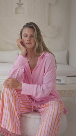 Woman wearing a pink shirt over an orange top and striped pants sitting on an ottoman in a neutral setting