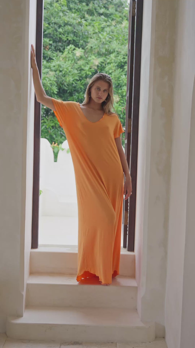 Woman in an orange maxi dress standing in a stylish room in front of a window.