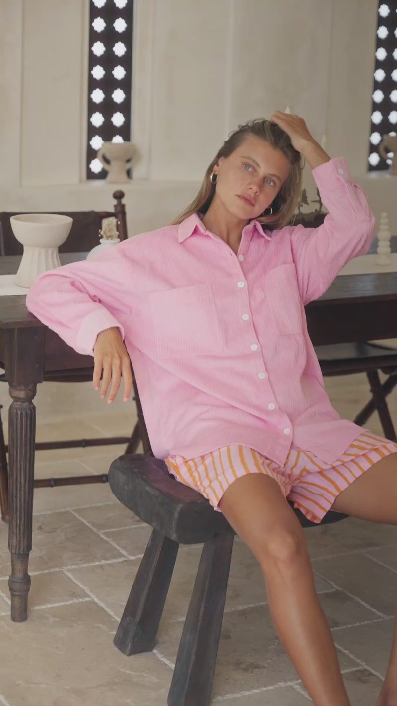Woman wearing a pink corduroy shirt  and striped shorts, sitting on a white surface.