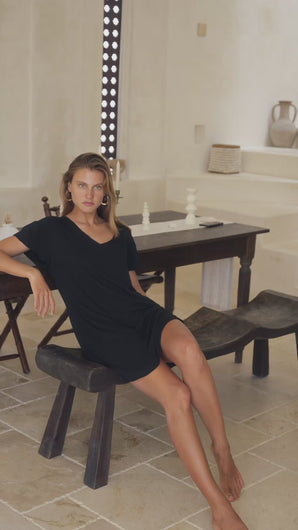 Woman in a black mini dress sitting on a wooden stool in a room with a mirror and decorative items.