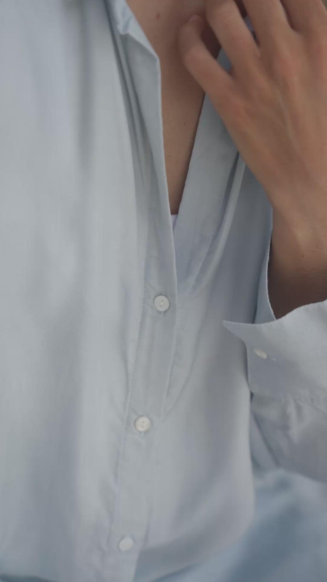 Load video: Woman wearing a light blue shirt over  a white tank top sitting against a neutral wall.