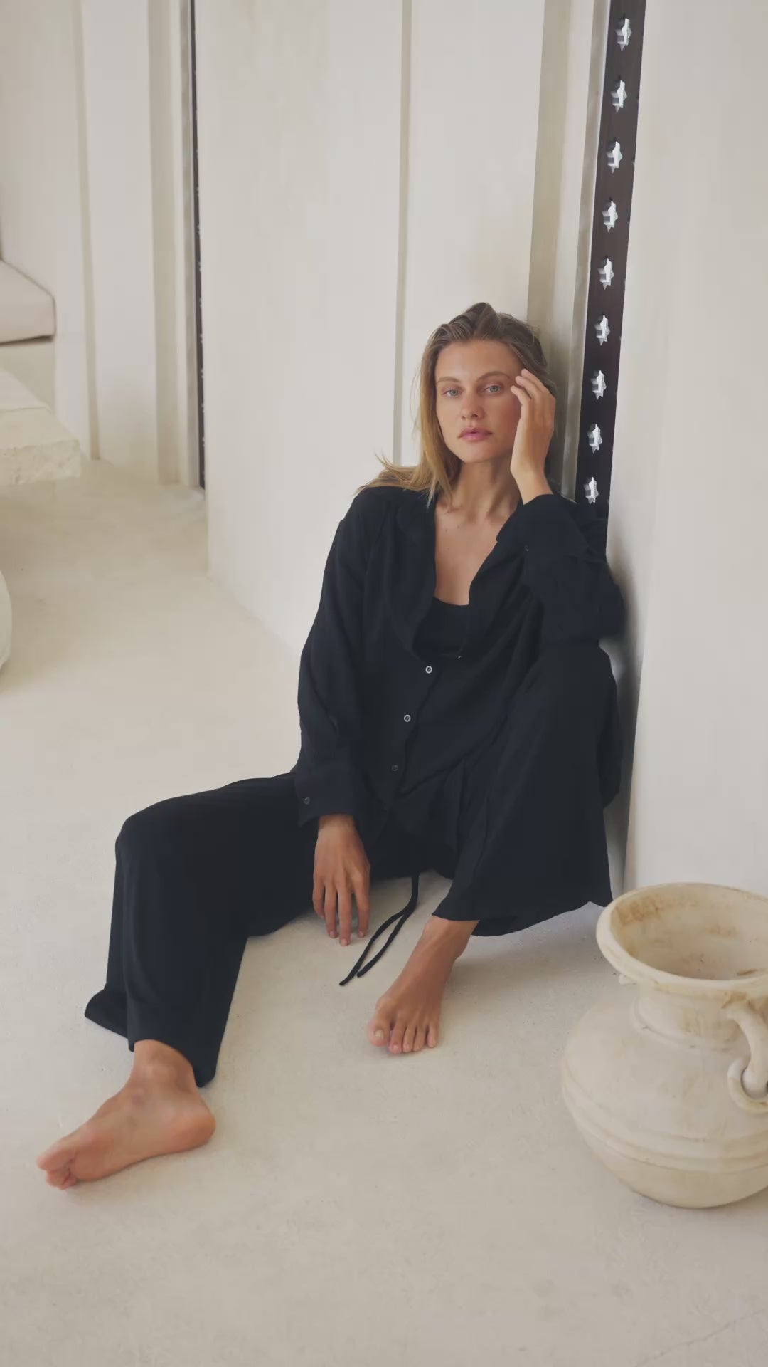 Load video: Woman wearing a black shirt, black pants and black tank top sitting on a white floor with a vase in the background