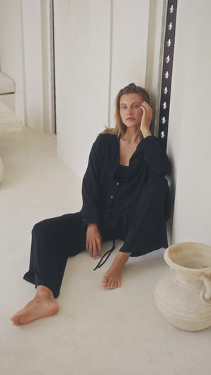 Woman wearing a black shirt, black pants and black tank top sitting on a white floor with a vase in the background