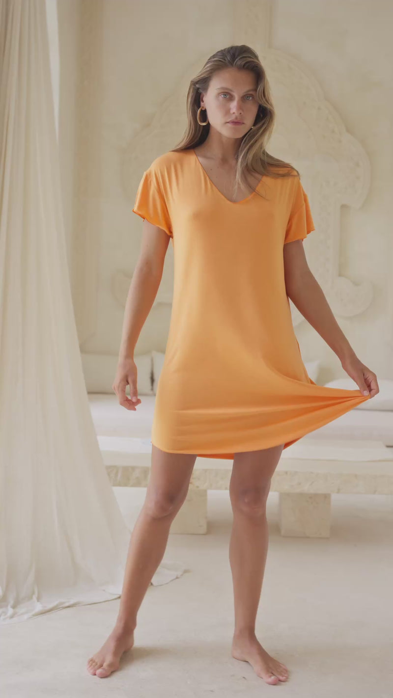 Woman wearing an orange mini dress standing near flowing curtains.