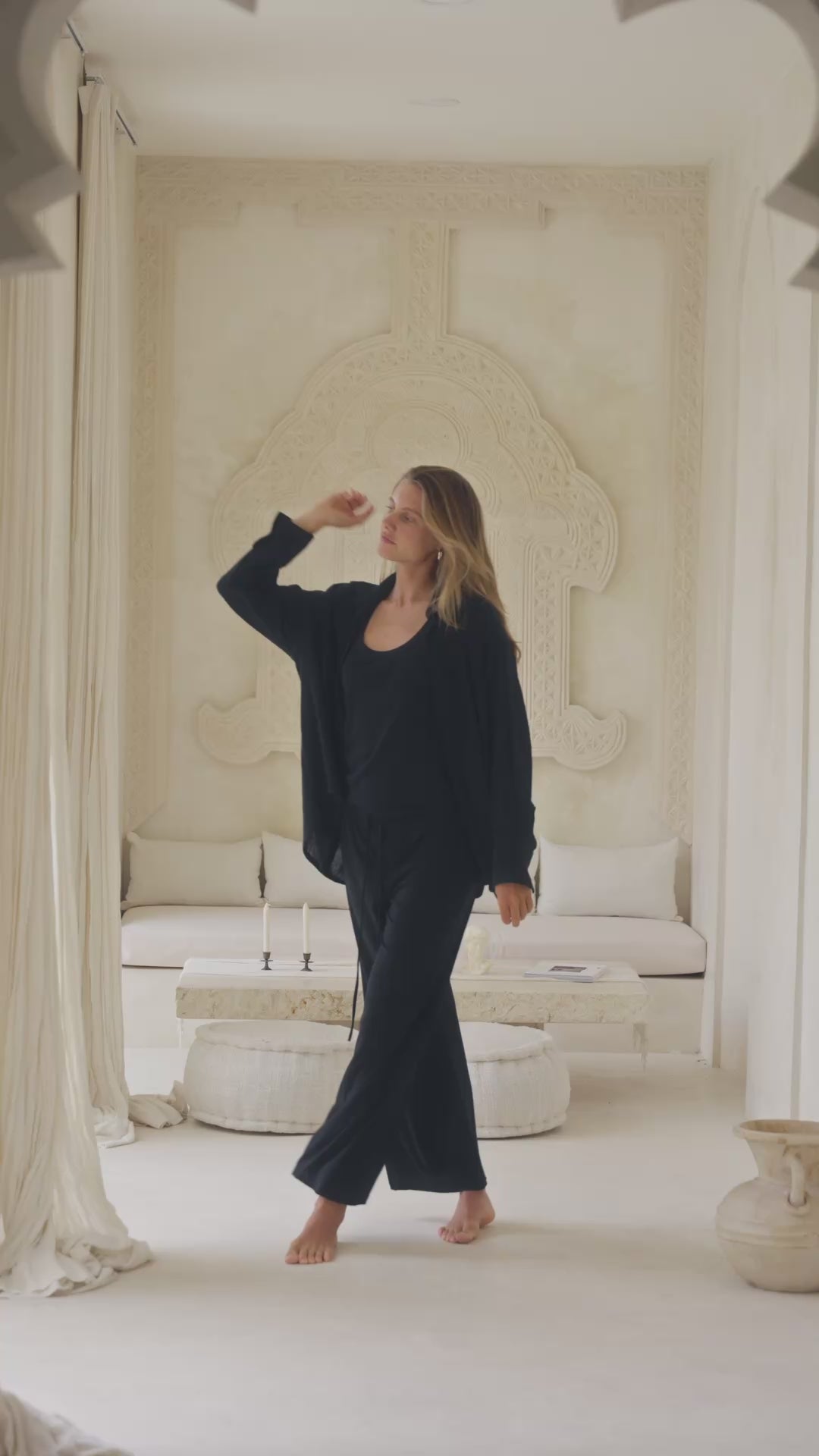 Load video: Woman in a black shirt, tank top and pants standing in a luxurious room with white decor