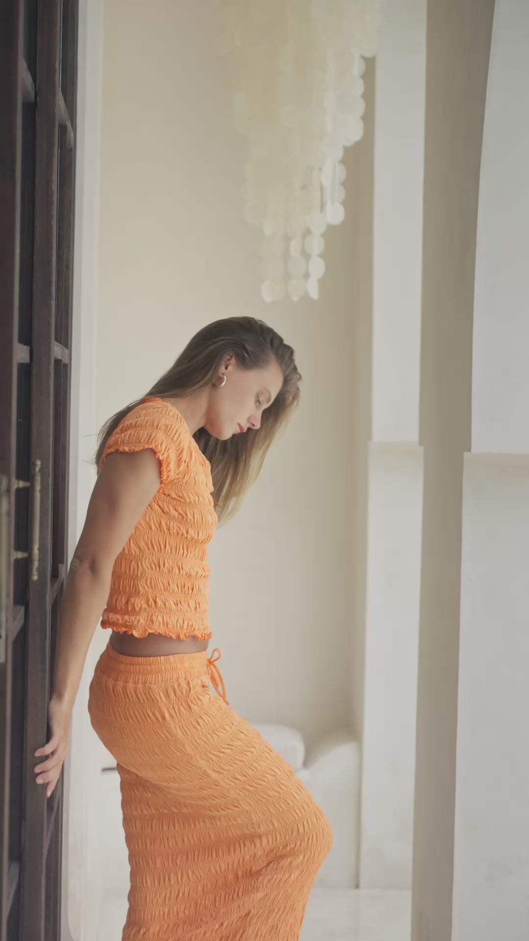 Load video: Woman in orange shirred top and maxi skirt standing on steps with a decorative wall in the background