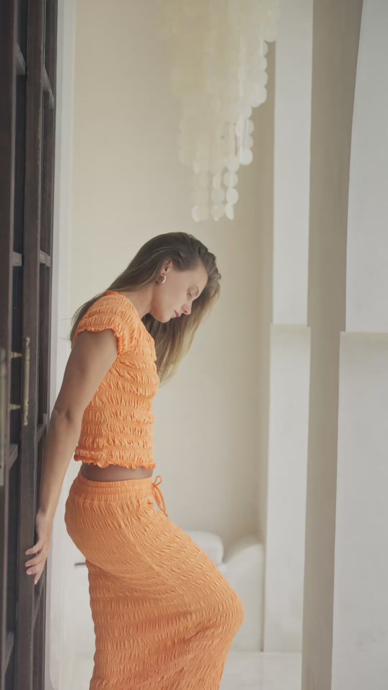 Woman in orange shirred top and maxi skirt standing on steps with a decorative wall in the background